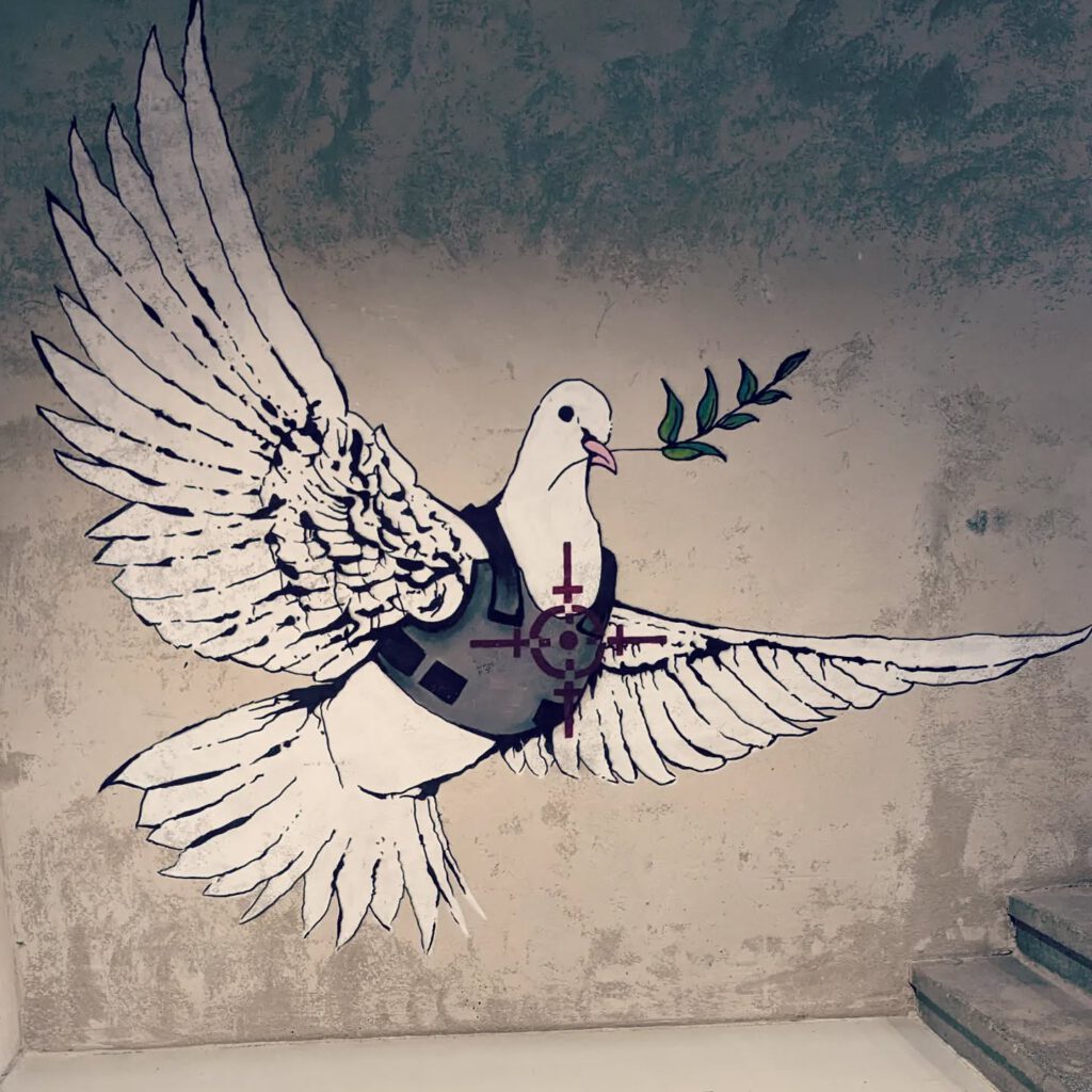 eine Friedenstaube mit Zweig im Schnabel trägt eine Schußweste. Auf die Taube ist ein Fadenkreuz gerichtet.