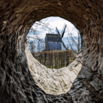 Das Foto wurde durch ein Loch aufgenommen, welches in einen dicken Baumstamm gehauen worden ist. Man sieht die Spuren der Bearbeitung. Durch das Loch fotografiert worden ist eine Bockwindmühle in einem Freilichtmuseum. Es scheint die Sonne.