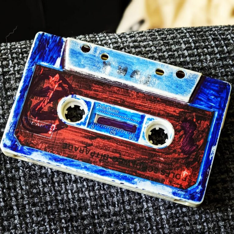 eine alte Musikkassette mit roten Ettikett wurde mit einem blauen Edding übermalt. Die Kassette sieht sehr alt aus. man sieht unter dem blauen Gekritzel den Schriftzug "Rolfs Schulweg Hitparade"