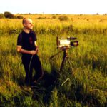 Ein junger Mann mit Glatze steht lachend auf einem Feld in der Abendsonne. Vor ihm steht eine Filmkamera