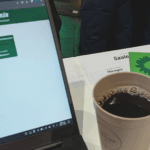 links ist ein aufgeklappter Laptop mit einer webseite der GRÜNEN zu sehen. Daneben steht ein Kaffeebecher. Im Hintergrund ist ein Ausschnitt eines Schildes mit dem Wort "Saale" zu sehen