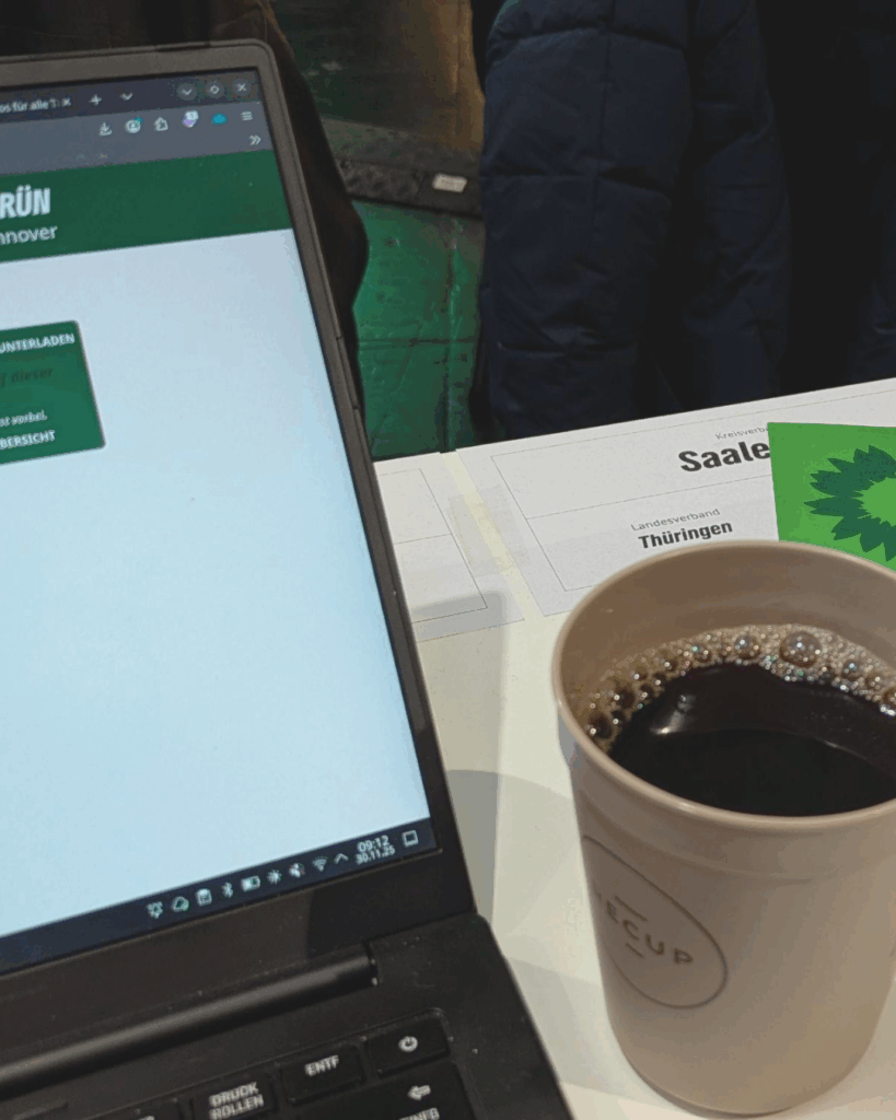 links ist ein aufgeklappter Laptop mit einer webseite der GRÜNEN zu sehen. Daneben steht ein Kaffeebecher. Im Hintergrund ist ein Ausschnitt eines Schildes mit dem Wort "Saale" zu sehen