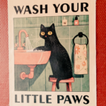 an einer Wand hängt ein Bild. Darauf sieht man eine gezeichnete schwarze Katze, die sich am Waschbecken die Pfoten wäscht. Dazu der Text: Wash you little Paws