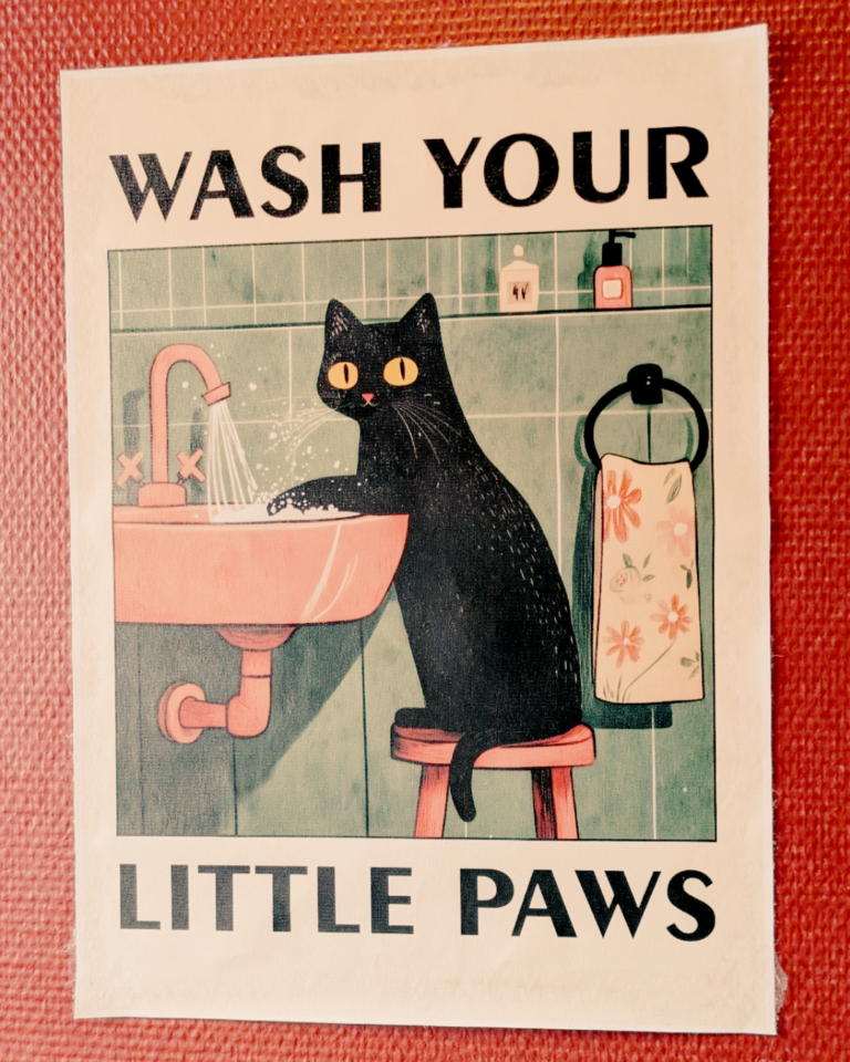 an einer Wand hängt ein Bild. Darauf sieht man eine gezeichnete schwarze Katze, die sich am Waschbecken die Pfoten wäscht. Dazu der Text: Wash you little Paws