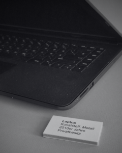 SchwarzWeiss-Foto von einem Laptop in einer Vitrine. Dazu ein kleines Schild mit dem Text: Laptop Kunststoff metall 2010er Jahre Privatbesitz