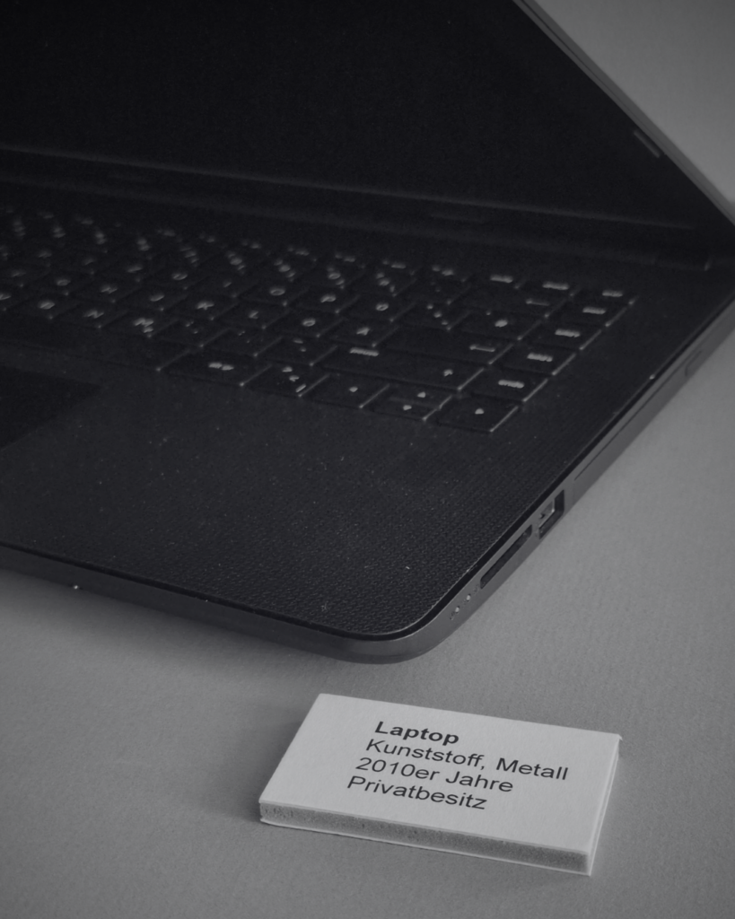 SchwarzWeiss-Foto von einem Laptop in einer Vitrine. Dazu ein kleines Schild mit dem Text: Laptop Kunststoff metall 2010er Jahre Privatbesitz